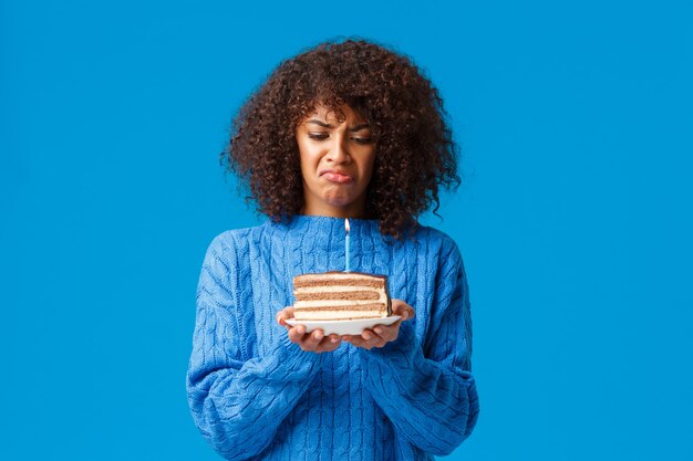 Boos en sombere, verontruste jonge Afro-Amerikaanse vrouw haat het vieren van verjaardag zich ouder voelen, lastig en ontevreden kijken naar verjaardagstaart met aangestoken kaars, mokkend, blauwe muur.