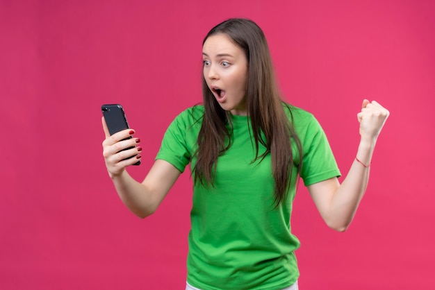 Boos jong mooi meisje dragen groene t-shirt met smartphone kijken scherm verbaasd en verrast staande over geïsoleerde roze achtergrond