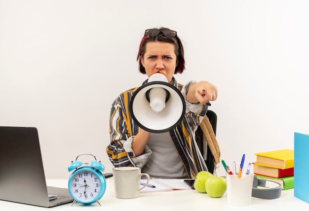 Gratis foto boos jong studentenmeisje die glazen op hoofd dragen die aan bureau zitten dat door spreker spreekt en op wit wordt geïsoleerd wijzen