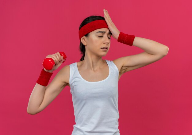 Boos jonge fitness vrouw in sportkleding met hoofdband halter in opgeheven hand houden op zoek verward met hand op haar hoofd staande over roze muur