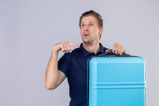 Boos jonge knappe reiziger man met koffer wegkijken wijzende vinger naar zijn koffer staande op witte achtergrond