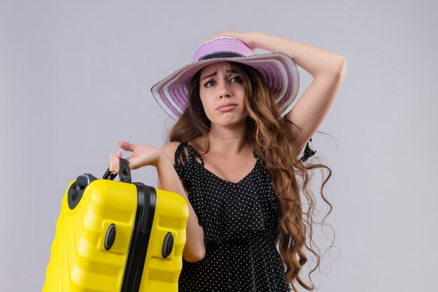 Boos jonge mooie reiziger meisje in jurk in polka dot in zomer hoed bedrijf koffer camera kijken met droevige uitdrukking op gezicht staande op witte achtergrond