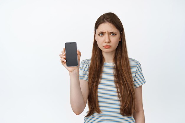 Boos jonge vrouw die haar telefoonscherm toont en teleurgesteld fronst Meisje houdt smartphone toont mobiel scherm met droevig boos gezicht staat tegen witte achtergrond