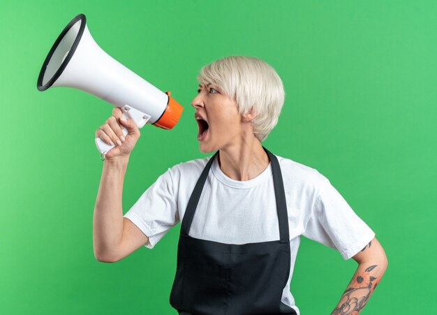 Boos kijken naar kant jonge mooie vrouwelijke kapper in uniform spreekt op luidspreker geïsoleerd op groene muur