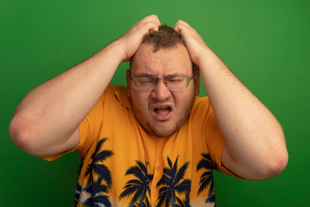 Gratis foto boos man in bril en oranje shirt op zoek geïrriteerd en geïrriteerd met handen op zijn hoofd staande over groene muur