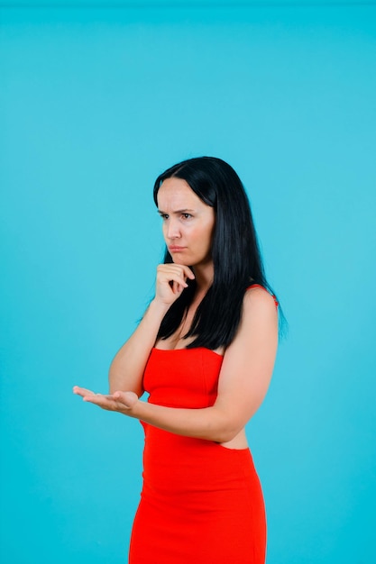 Boos meisje denkt door hand op kin te houden en andere naar links op blauwe achtergrond uit te breiden