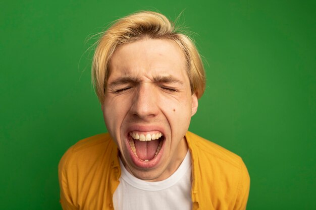 Boos met gesloten ogen jonge blonde kerel die gele t-shirt draagt die op groen wordt geïsoleerd