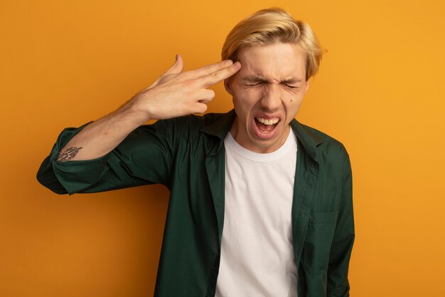 Gratis foto boos met gesloten ogen jonge blonde kerel die groen t-shirt draagt dat zelfmoord met pistoolgebaar toont