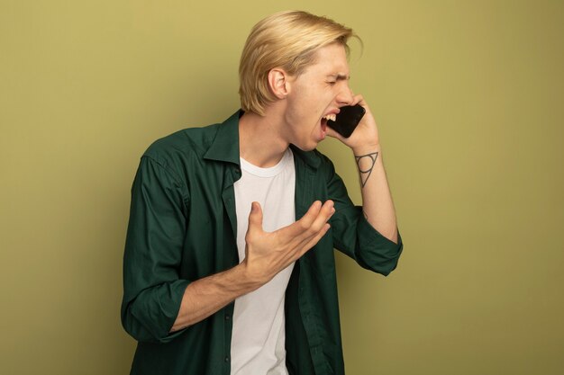 Boos met gesloten ogen spreekt de jonge blonde kerel die een groen t-shirt draagt aan de telefoon