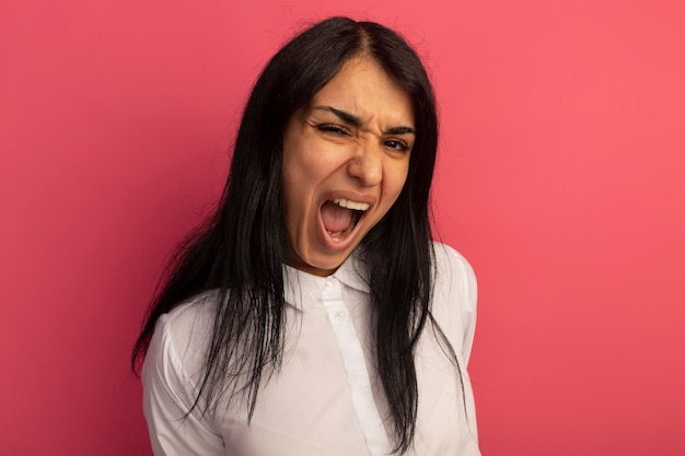 Boos recht vooruit kijkend jong mooi meisje dat wit t-shirt draagt dat op roze wordt geïsoleerd