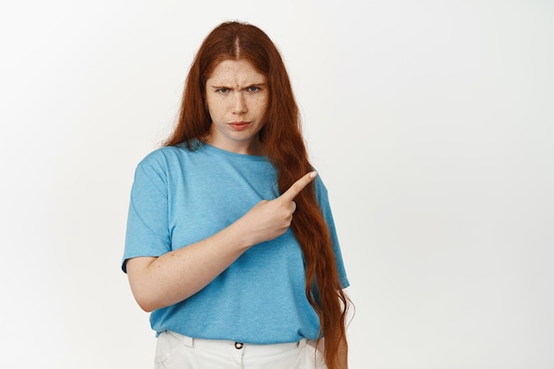 Boos roodharig tienermeisje, wenkbrauwen fronsen en wijzend naar de rechterbovenhoek met een ontevreden uspet-gezicht, klagen, iets slecht beoordelen, in t-shirt tegen witte achtergrond staan