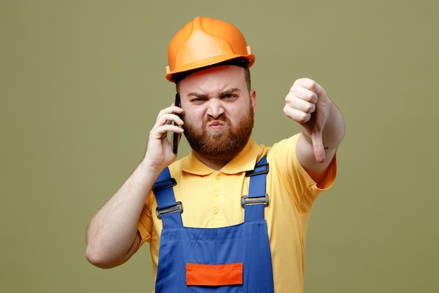 Gratis foto boos tonen duim omlaag spreekt op telefoon jonge bouwer man in uniform geïsoleerd op groene achtergrond