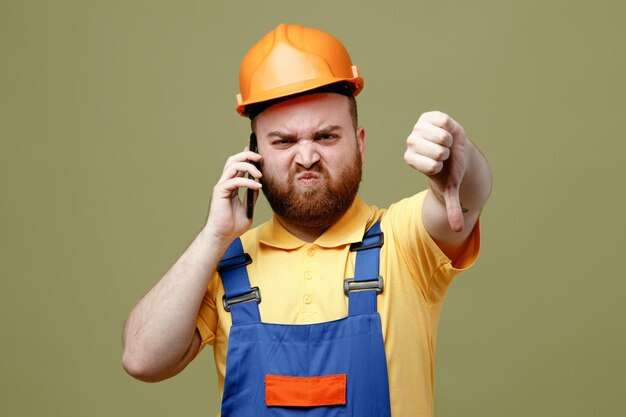 Boos tonen duim omlaag spreekt op telefoon jonge bouwer man in uniform geïsoleerd op groene achtergrond