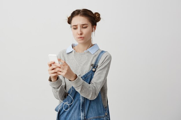 Boos trendy meisje op zoek op smartphone met spijt blik en samengeknepen lippen. Brunette vrouw kan favoriete muziek niet vinden in haar gadget die probeert te uploaden. Techniek concept