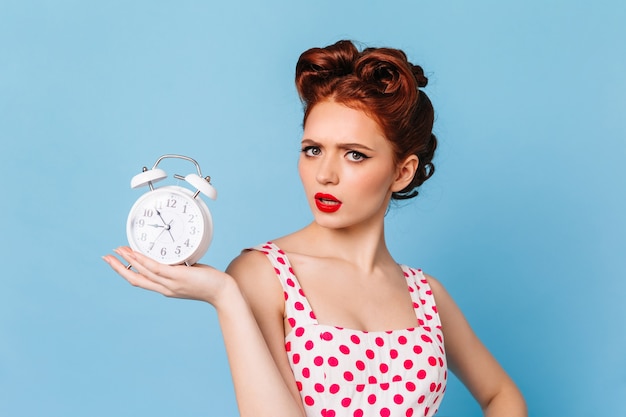 Boos vrouw met lichte make-up die tijd toont. Studio die van mooi pinupmeisje met klok is ontsproten.