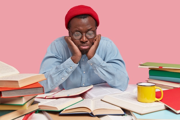 Boos zwarte jongeman gefocust naar beneden, voelt zich depressief en verdrietig terwijl hij lange tijd boeken leest, ronde bril, rode hoed en shirt draagt