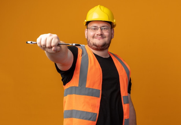 Bouwer in bouwvest en veiligheidshelm met moersleutel die naar de camera kijkt met een zelfverzekerde glimlach op het gezicht over oranje achtergrond