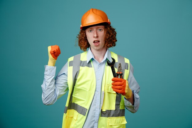 Bouwer jonge man in bouw uniform en veiligheidshelm met moersleutel en tang balde vuist blij en opgewonden permanent over blauwe achtergrond