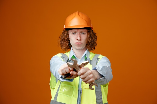 Bouwer jonge man in bouw uniform en veiligheidshelm met moersleutels die handen kruisen kijkend naar camera met fronsend gezicht doen stop gebaar staande over oranje achtergrond