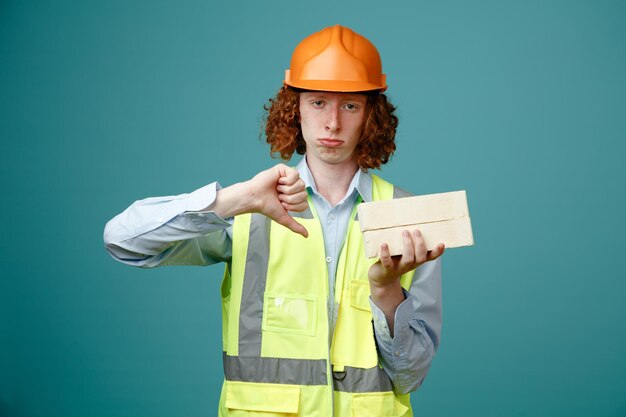 Bouwer jonge man in bouw uniform en veiligheidshelm met twee bakstenen kijken camera ontevreden tonen duim naar beneden staande over blauwe achtergrond