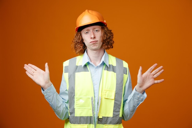 Bouwer jonge man in bouwuniform en veiligheidshelm kijkend naar camera met sceptische uitdrukking die armen opheft over oranje achtergrond