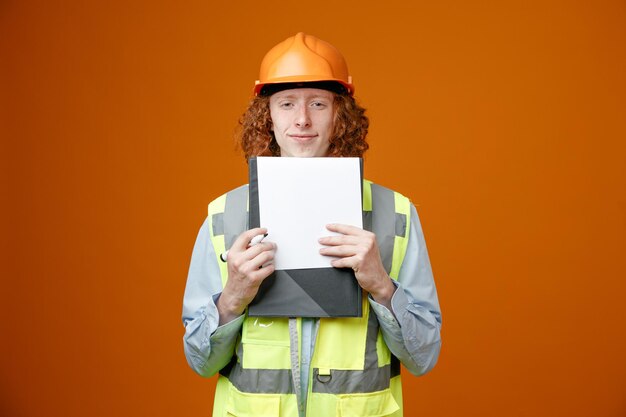 Bouwer jonge man in bouwuniform en veiligheidshelm met klembord en marker kijkend naar camera glimlachend zelfverzekerd over oranje achtergrond
