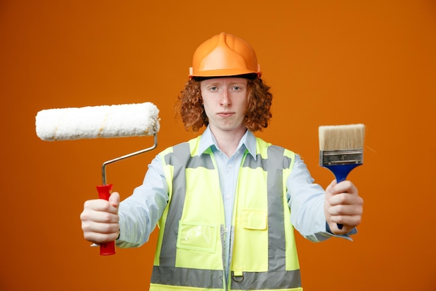 Bouwer jonge man in bouwuniform en veiligheidshelm met verfroller en penseel kijkend naar camera met serieus gezicht over oranje achtergrond
