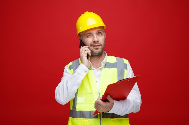 Bouwer man in bouw uniform en veiligheidshelm met klembord praten op mobiele telefoon glimlachend met sceptische uitdrukking staande over rode achtergrond