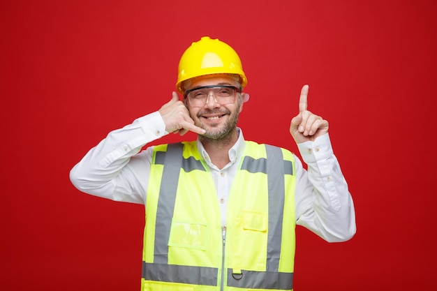 Bouwer man in bouw uniform en veiligheidshelm met veiligheidsbril kijkend naar camera glimlachend vrolijk maken bel me gebaar wijzend met wijsvinger omhoog permanent over rode achtergrond