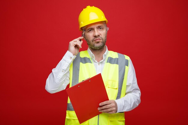 Bouwer man in constructie uniform en veiligheidshelm met klembord kijkend naar camera verbaasd krabben zijn hoofd staande over rode achtergrond