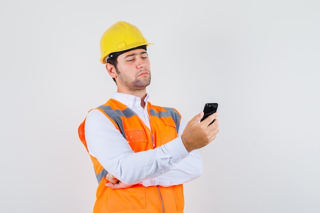 Bouwer man kijken naar smartphone in shirt, uniform en peinzend, vooraanzicht kijken.