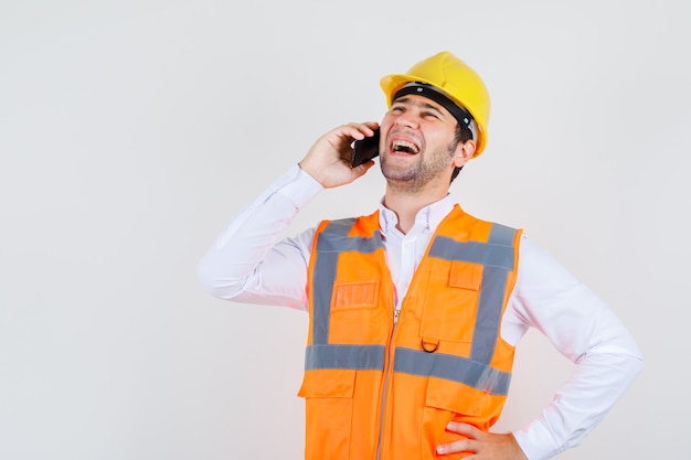 Bouwer man praten over smartphone met hand op taille in shirt, uniform en op zoek vrolijk. vooraanzicht.