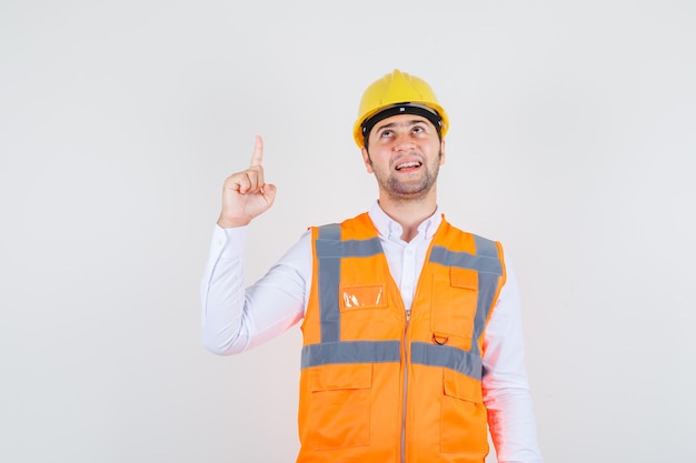 Bouwer man wijzende vinger in shirt, uniform en op zoek vrolijk. vooraanzicht.