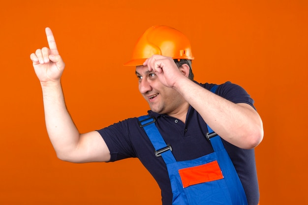 Bouwersmens die eenvormige bouw dragen en veiligheidshelm die zich met gelukkig gezicht bevinden en wijzende vinger omhoog en wat betreft zijn helm over geïsoleerde oranje muur glimlachen