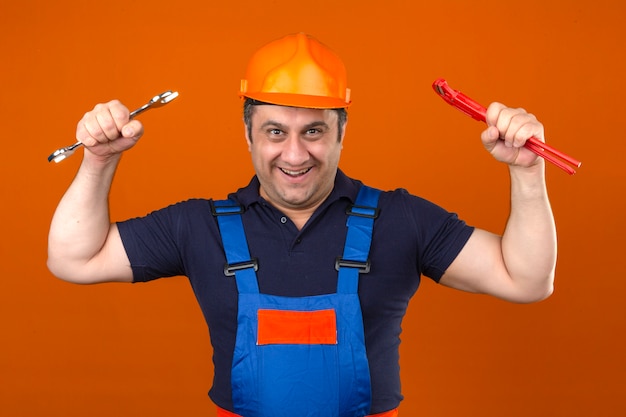 Gratis foto bouwersmens die eenvormige bouw dragen en veiligheidshelm die zich met moersleutels in opgeheven handen bevinden met gelukkig gezicht over geïsoleerde oranje muur