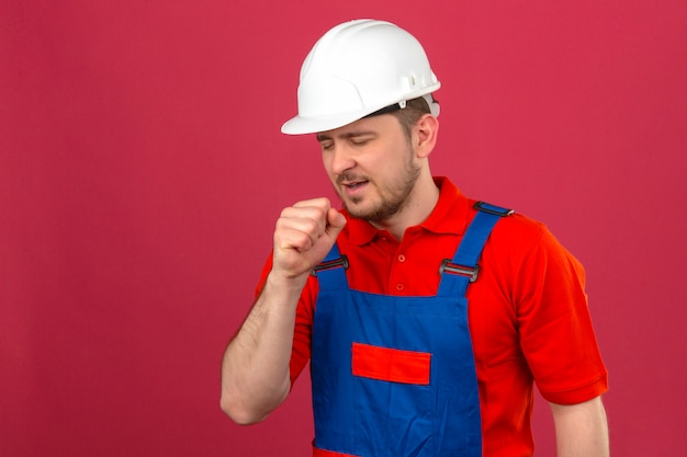 Bouwersmens die eenvormige bouw dragen en veiligheidshelm die zich onwel voelen en hoesten die zich over geïsoleerde roze muur bevinden