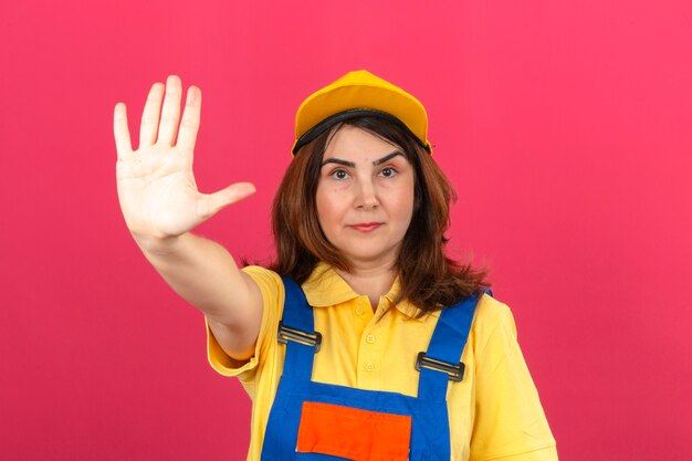Bouwersvrouw die bouw eenvormig en geel GLB dragen die zich met open hand bevinden die eindeteken met het ernstige en zekere gebaar van de uitdrukkingsdefensie doen over geïsoleerde roze muur