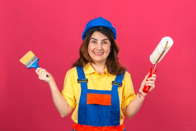 Bouwersvrouw eenvormige bouw dragen en de holdingsborstel van de veiligheidshelm en verfrol in handen die vrolijk over geïsoleerde roze muur glimlachen