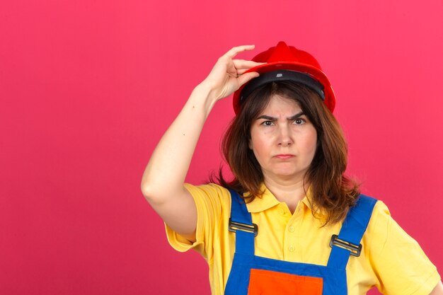 Bouwersvrouw in eenvormige bouw en veiligheidshelm met het fronsen gezicht wat betreft helm met hand die zich over geïsoleerde roze muur bevinden