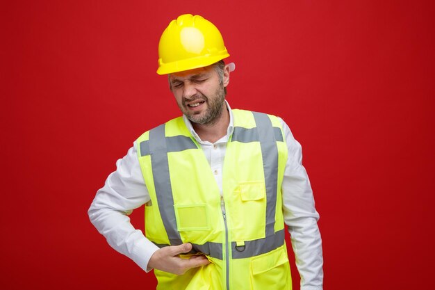Bouwman in bouwuniform in veiligheidshelm die er onwel uitziet en zijn buik aanraakt, voelt pijn die over rode achtergrond staat