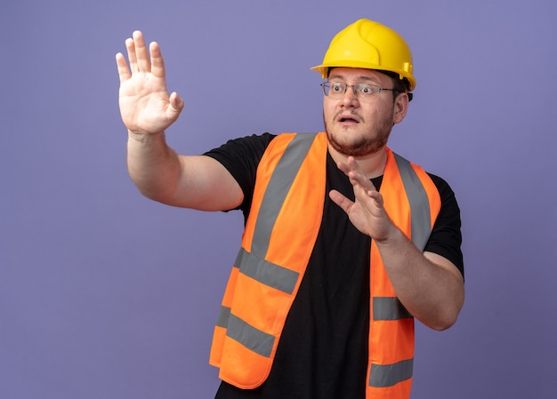 Bouwman in bouwvest en veiligheidshelm kijkt opzij, bezorgd hand in hand terwijl hij over blauw staat