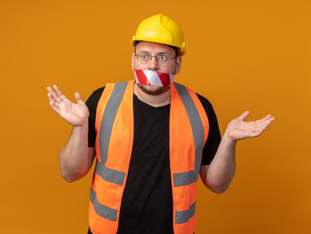 Bouwman in bouwvest en veiligheidshelm met tape over zijn mond die er verward uitziet en armen naar de zijkanten spreidt