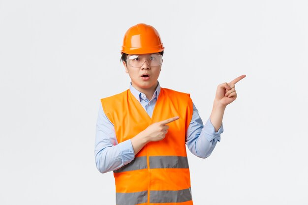 Bouwsector en industriële werknemers concept. Onder de indruk en verbaasd Aziatische bouwmanager, ingenieur in helm en reflecterende kleding, wijzend op de rechterbovenhoek, witte muur
