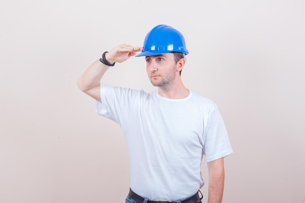 Bouwvakker in t-shirt, spijkerbroek, helm die een groetgebaar toont en er gefocust uitziet