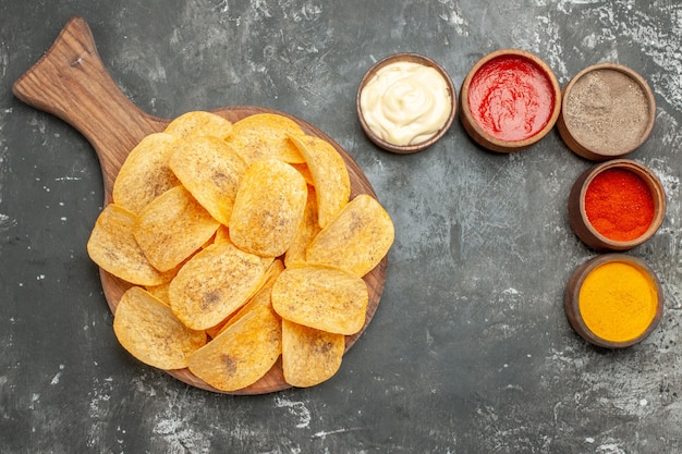 Boven weergave van aardappelchips, kruiden en mayonaise met ketchup op houten snijplank op grijze tafel
