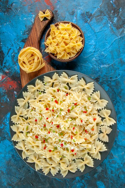 Boven weergave van farfalle pasta's met groenten op een zwarte plaat op houten snijplank en verschillende pasta's op blauwe achtergrond