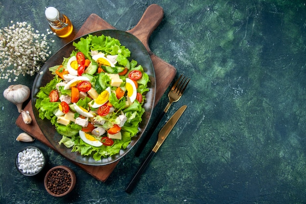 Boven weergave van heerlijke salade met veel verse ingrediënten op houten snijden boa rd specerijen oliefles bestek aan de rechterkant op zwarte groene mix kleuren tafel