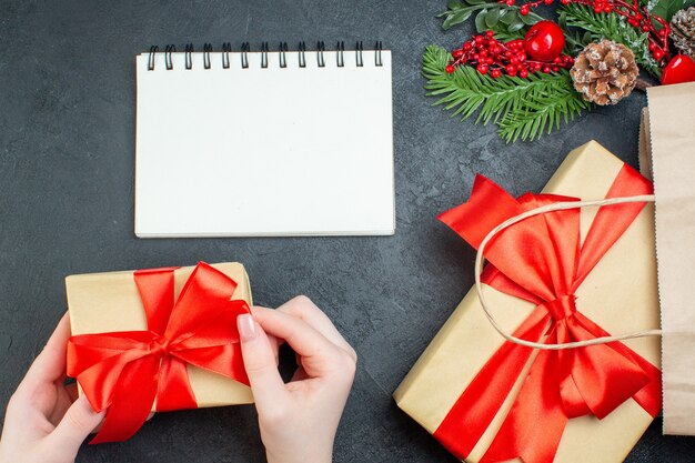 Boven weergave van kerstsfeer met hand met een van de mooie geschenken en fir takken conifer kegel naast notebook op donkere achtergrond