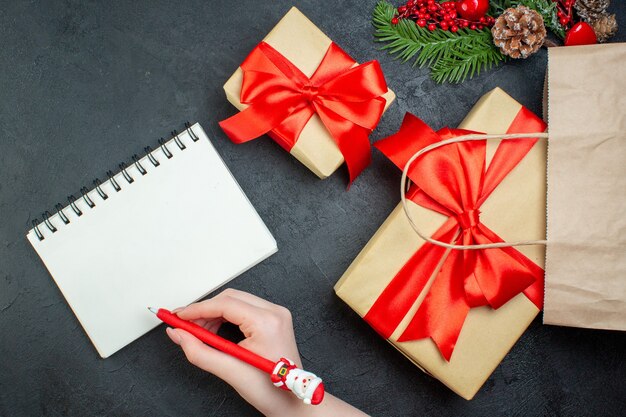 Boven weergave van kerstsfeer met mooie geschenken en fir takken conifer kegel naast notebook met pen op donkere achtergrond