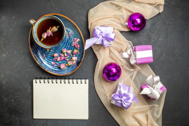 Boven weergave van kleurrijke geschenken decoratie accessoires voor Kerstmis op naakt kleur handdoek en een kopje thee naast notebook op zwarte achtergrond